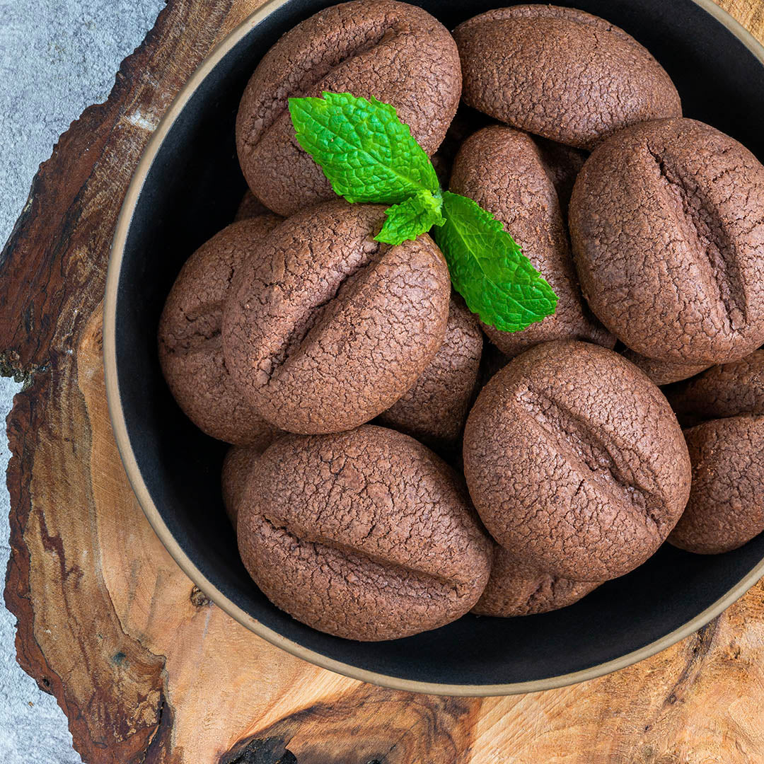 A bowl of chocolate cookies shaped like coffee beans, sweetened with Roasted Espresso Monk Fruit Sweetener and garnished with fresh mint leaves, sits on a wooden surface.