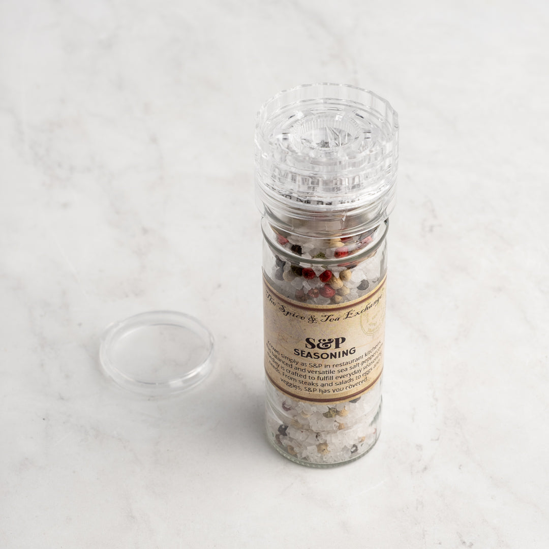 A clear plastic S & P Seasoning grinder, filled with mixed peppercorns and sea salt, sits on a light marble surface with its lid removed and placed beside it.