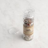 A clear plastic S & P Seasoning grinder, filled with mixed peppercorns and sea salt, sits on a light marble surface with its lid removed and placed beside it.