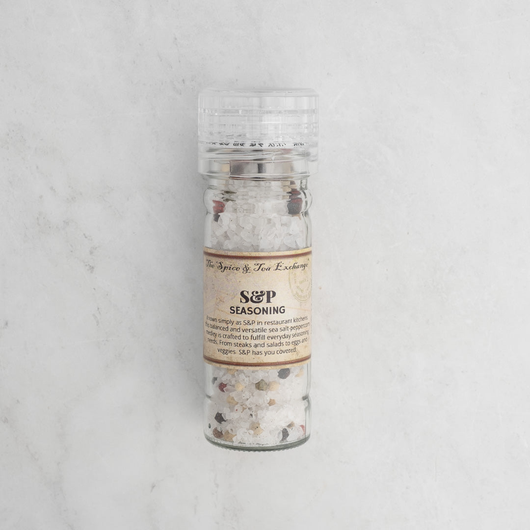 A clear plastic grinder of S & P Seasoning, filled with a blend of sea salt and whole peppercorns, sits on a light textured marble surface.