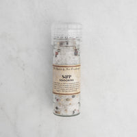 A clear plastic grinder of S & P Seasoning, filled with a blend of sea salt and whole peppercorns, sits on a light textured marble surface.
