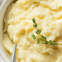 Close-up of creamy mashed potatoes in a white bowl, garnished with a sprig of fresh thyme and a silver spoon visible at the lower left.