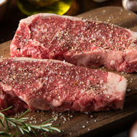 Two raw beef steaks seasoned with coarse sea salt and black pepper, resting on a dark wooden cutting board with rosemary sprigs and a blurred olive oil bottle in the background.