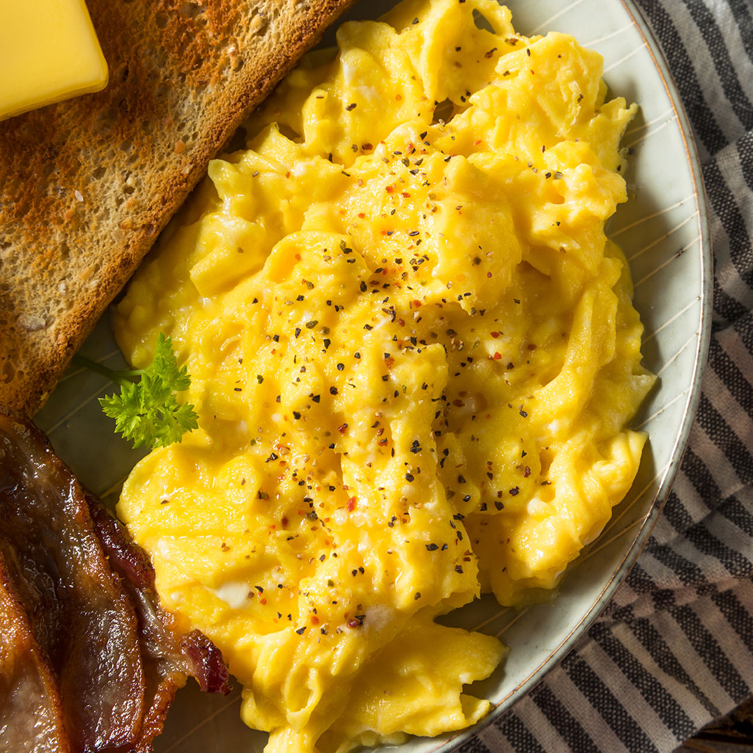 A close-up overhead shot of a breakfast plate featuring fluffy scrambled eggs topped with black pepper and a sprinkle of red pepper flakes, served with a slice of toast, bacon, and a small sprig of parsley on a striped plate.