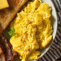 A close-up overhead shot of a breakfast plate featuring fluffy scrambled eggs topped with black pepper and a sprinkle of red pepper flakes, served with a slice of toast, bacon, and a small sprig of parsley on a striped plate.
