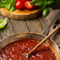 A close-up of a rustic metal pan filled with rich tomato sauce, with a wooden spoon resting inside. Fresh basil leaves and sliced tomatoes are visible in the softly blurred background on a dark wooden table.
