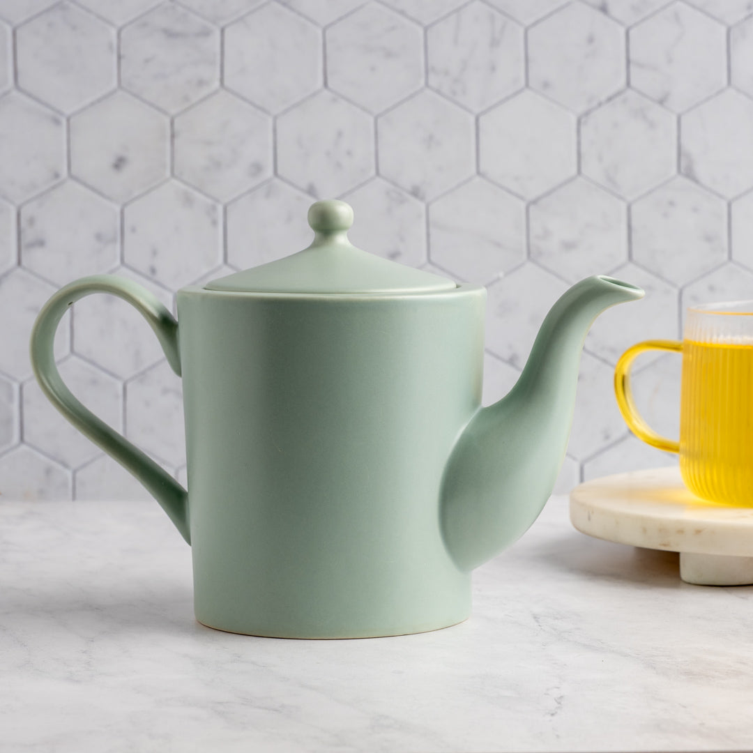 A light sage green ceramic teapot with a matching lid sits on a white marble surface in front of a white hexagonal tile backsplash. A yellow glass mug on a marble coaster is partially visible on the right.