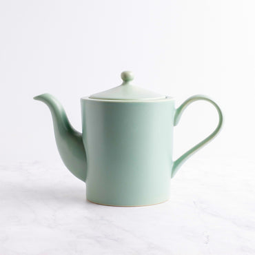 A light sage green ceramic teapot with a matching lid, resting on a white marble surface against a white background.