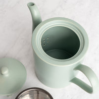 A light sage green ceramic teapot with a matching lid, shown on a white marble surface with a metal strainer visible at the bottom.