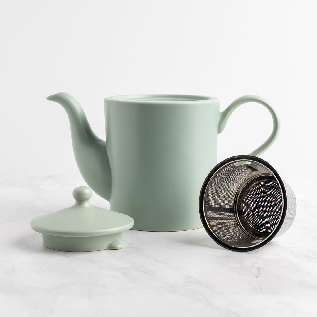 A light sage green ceramic teapot with a matching lid, shown on a light marble surface with a metal infuser filter resting beside it.