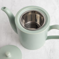 A light sage green ceramic teapot with a stainless steel mesh infuser inside, shown on a light marble surface with its matching lid partially visible in the lower left corner.
