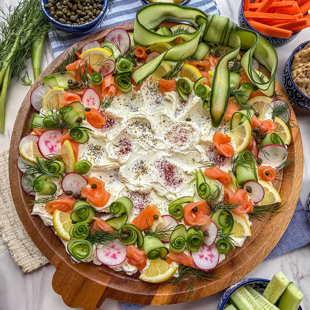 A large round wooden platter filled with a gourmet appetizer spread, featuring a thick layer of cream cheese, topped with rolled cucumber, sliced radishes, smoked salmon, lemon wedges, dill, and seasonings, surrounded by side bowls of capers, carrots, and cucumber sticks.