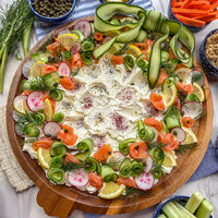 A large round wooden platter filled with a gourmet appetizer spread, featuring a thick layer of cream cheese, topped with rolled cucumber, sliced radishes, smoked salmon, lemon wedges, dill, and seasonings, surrounded by side bowls of capers, carrots, and cucumber sticks.