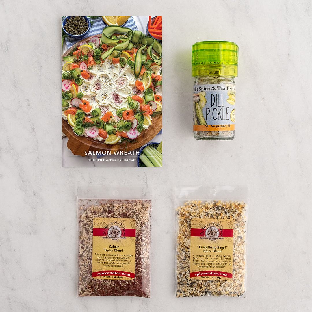 A flat-lay product arrangement on a light marble surface featuring a Salmon Wreath recipe card and 3 spice blends/seasonings.