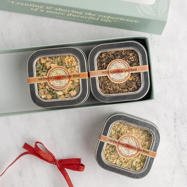 A gift set of two square metal tins of spice blends, shown with their matching light green presentation box and a red ribbon on a white marble surface. The tins feature circular labels.