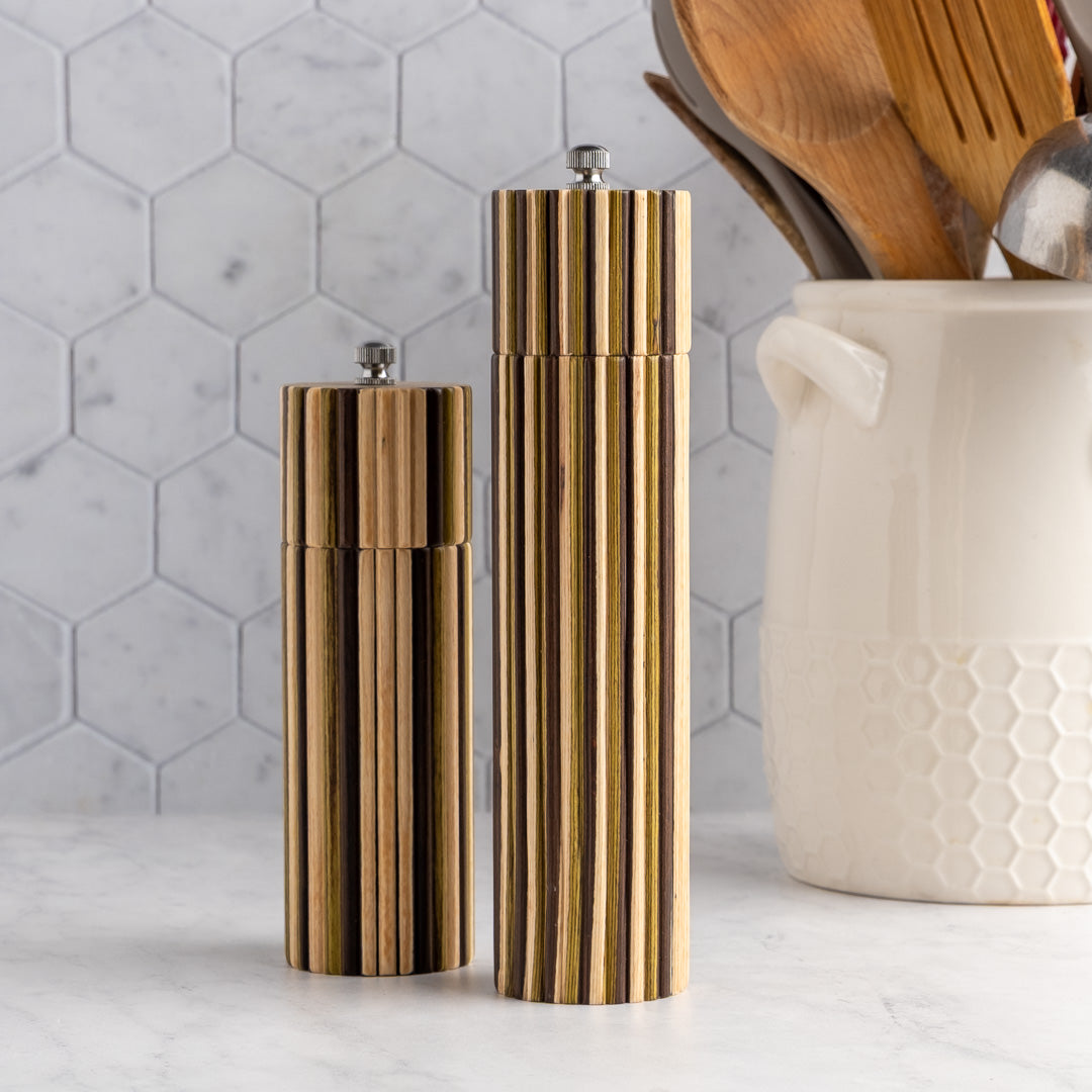 Two modern cylindrical salt and pepper mills with vertical fluted wood grain in warm brown and gold tones, standing on a light marble countertop against a white hexagonal tile backsplash. A white ceramic jar with wooden utensils sits to the right.