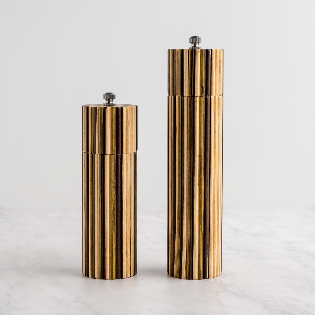 Two cylindrical striped salt and pepper mills with a gold and black wood-grain pattern, standing on a light marble surface against a white background.