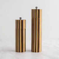Two cylindrical striped salt and pepper mills with a gold and black wood-grain pattern, standing on a light marble surface against a white background.