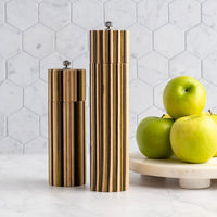 Two modern cylindrical salt and pepper mills with vertical brushed gold metal stripes and silver tops, displayed on a white marble surface next to green apples on a marble pedestal against a white hexagonal tile background.