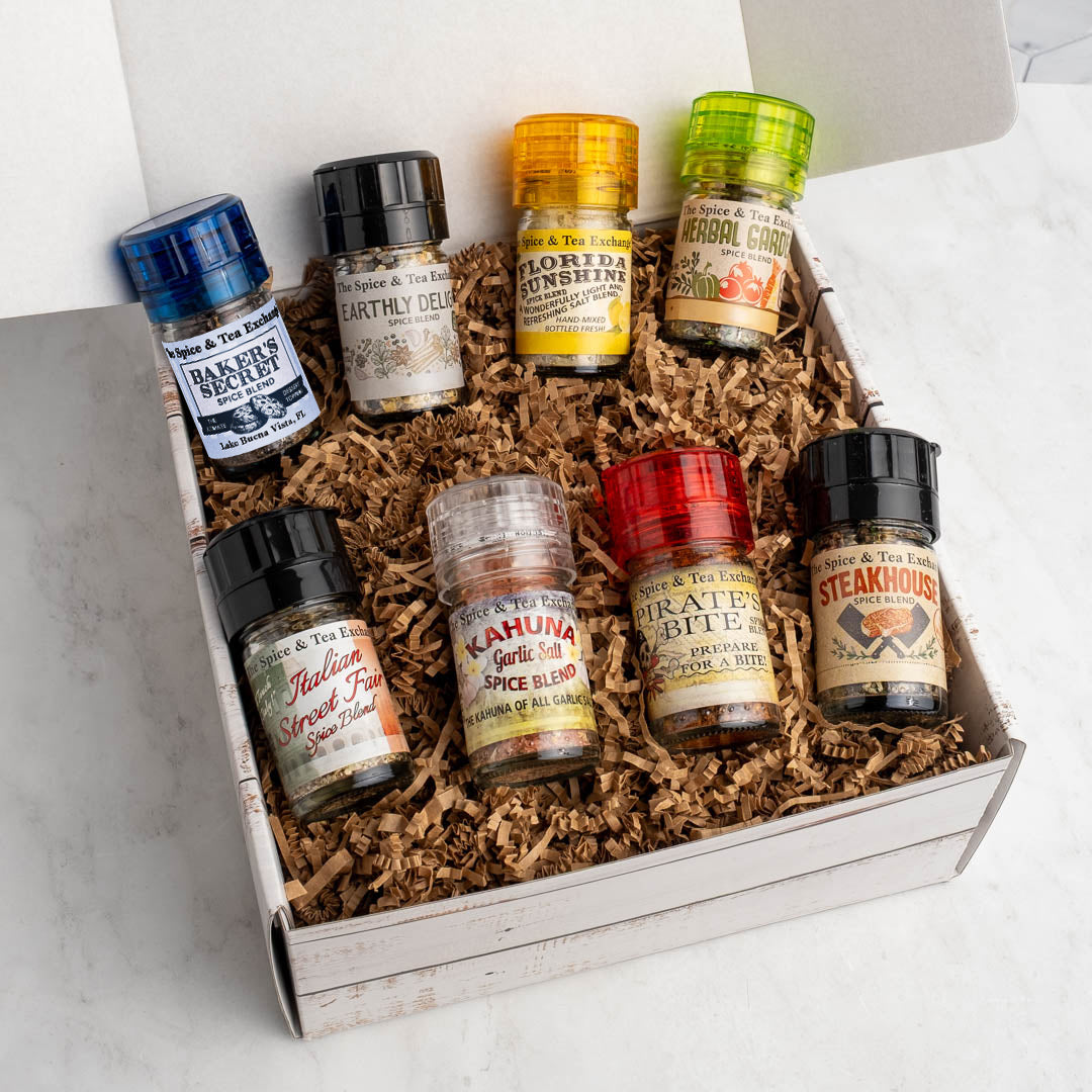 The Season Up! Deluxe Gift Box features eight assorted spice blend jars, each with a colorful lid and unique label, nestled in brown shredded paper—an ideal seasoning sampler—beautifully displayed on a white marble surface.