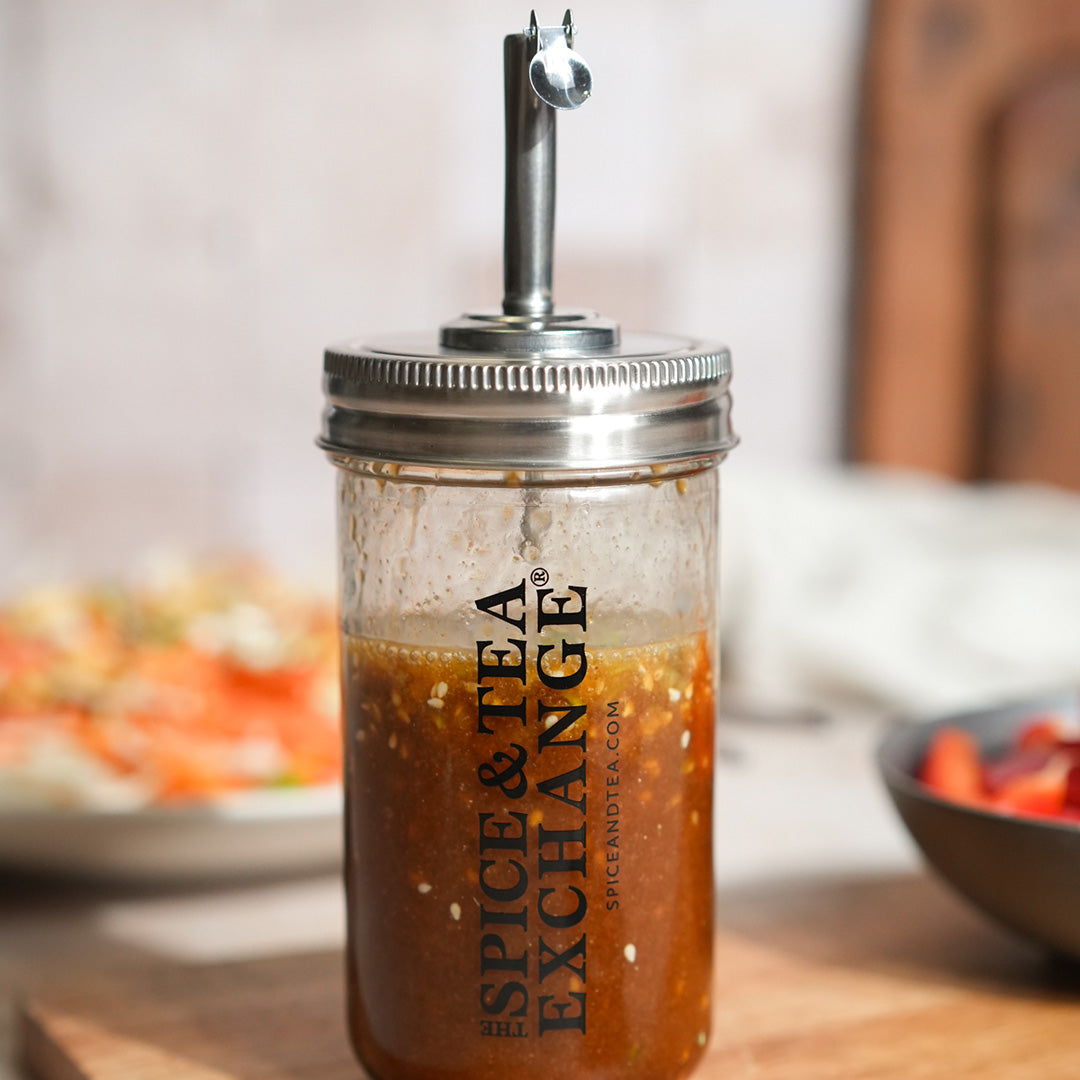 The Shake Strain & Pour Jar, featuring a metal pour spout and The Spice & Tea Exchange label, is filled with brown vinaigrette dressing and rests on a wooden surface with blurred dishes in the background.
