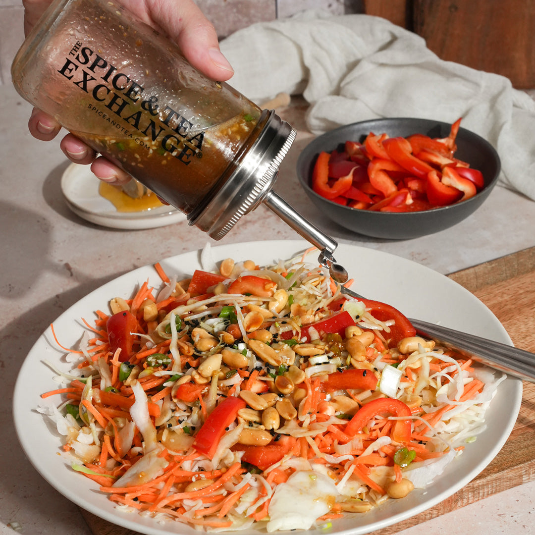 A hand pours dressing from the Shake Strain & Pour Jar onto a salad of shredded cabbage, carrots, red bell peppers, and peanuts, with extra sliced peppers in a bowl nearby.