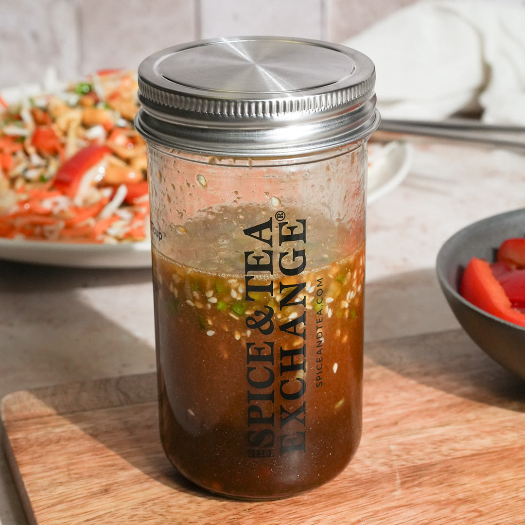 The Shake Strain & Pour Jar, a glass mixing jar with a metal lid labeled The Spice & Tea Exchange, holds a brown, speckled liquid and sits on wood amid salads—perfect for shaking and pouring salad dressings.