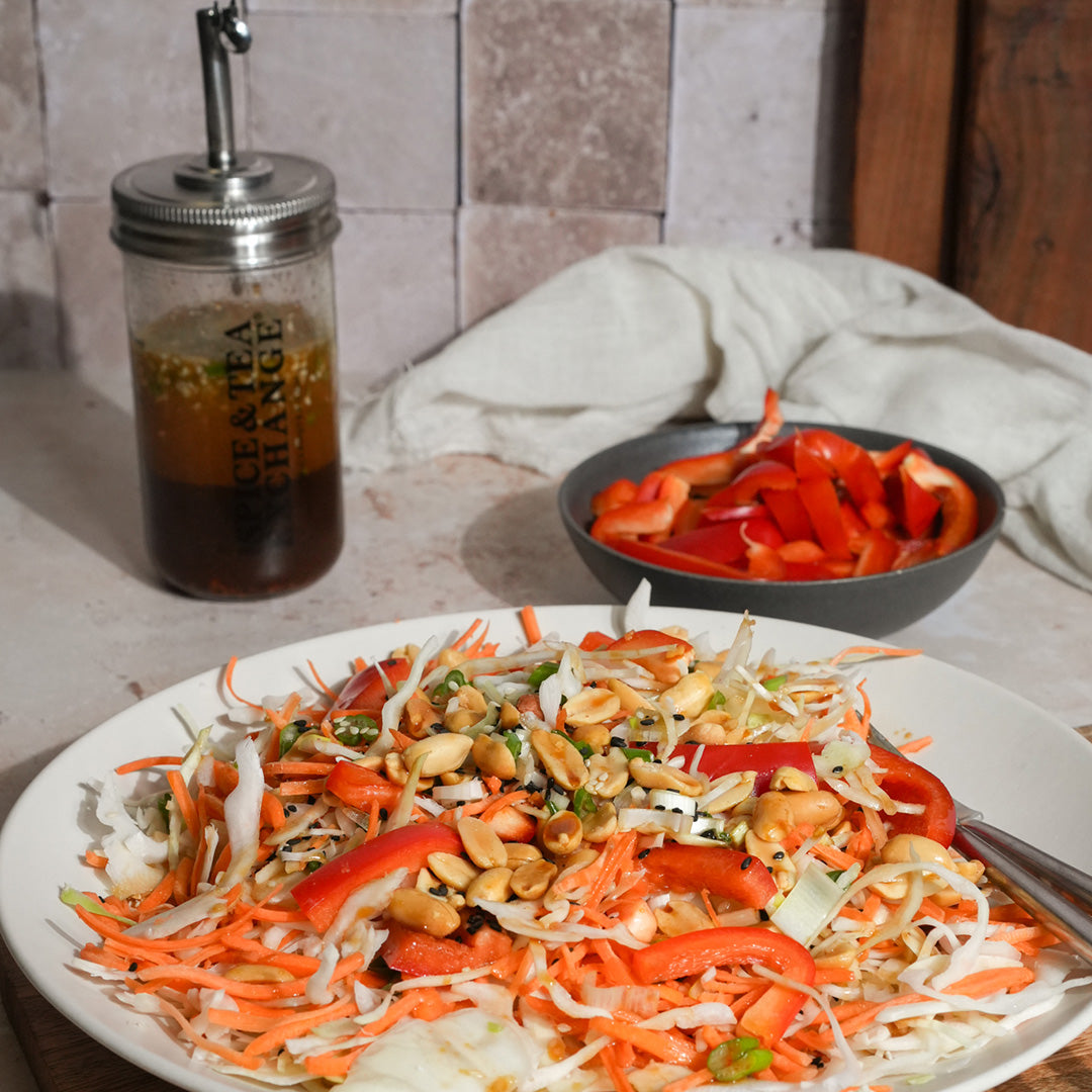 A plate of shredded cabbage, carrots, red bell peppers, and peanuts sits on a table. Behind it are a bowl of red bell peppers and the Shake Strain & Pour Jar. A white cloth is in the background.