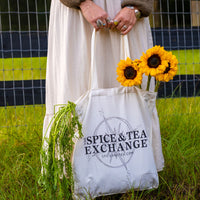 Person holding a tote bag with 'The Spice & Tea Exchange' logo, surrounded by sunflowers and greenery.