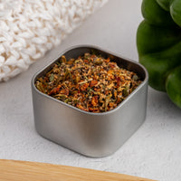 A square metal tin of Signature Spice Blend rests on a light surface beside a green bell pepper and a white woven item, capturing the vibrant flavors inspired by St. Augustine.