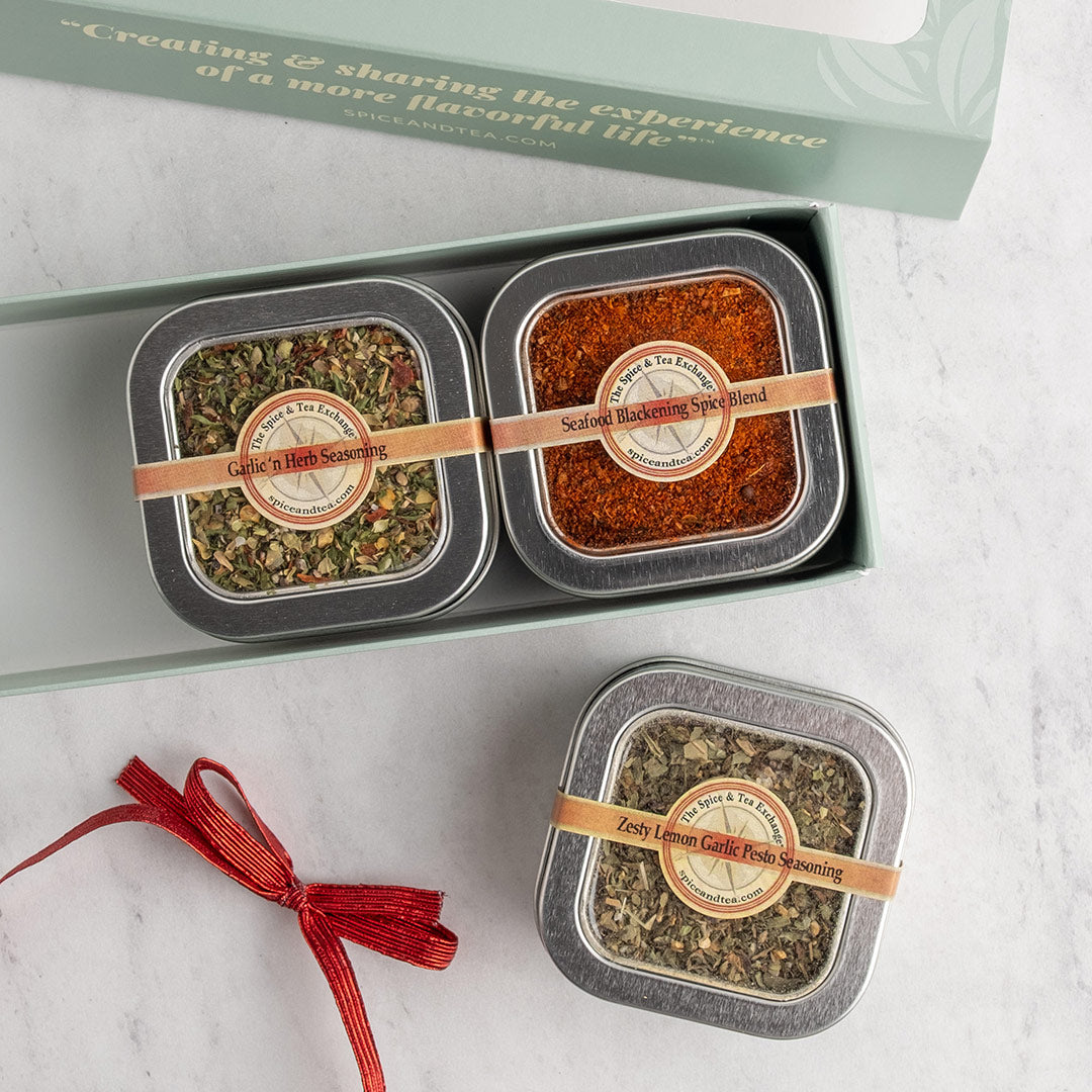 Three square metal tins of seasonings and spice blends arranged in and around a light green gift box on a white marble surface. A red ribbon is tied setting next to it.