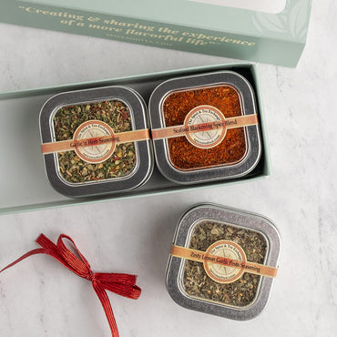 Three square metal tins of seasonings and spice blends arranged in and around a light green gift box on a white marble surface. A red ribbon is tied setting next to it.