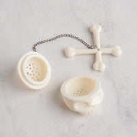 An off-white bone-themed tea infuser set on a light gray marble surface, featuring a skull-shaped strainer, a bone cross with a hanging bone charm, and a silver metal chain.
