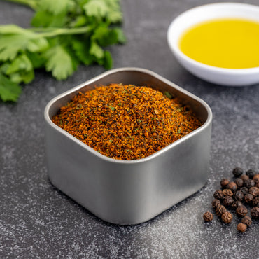 A metal tin of Smoky Black Garlic Seasoning rests on a dark countertop beside whole black peppercorns, fresh herbs, and a white bowl filled with yellow oil.
