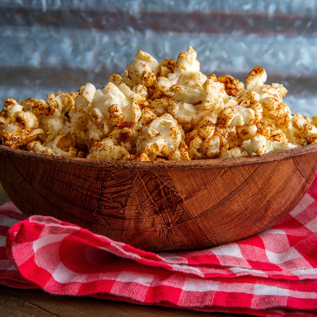 Seasoned popcorn piled in a carved wooden bowl resting on a red and white checkered cloth against a rustic blue-gray background.