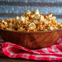 Seasoned popcorn piled in a carved wooden bowl resting on a red and white checkered cloth against a rustic blue-gray background.