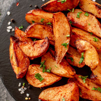 Crispy roasted potato wedges garnished with chopped green parsley, served on a dark slate platter with scattered coarse sea salt.