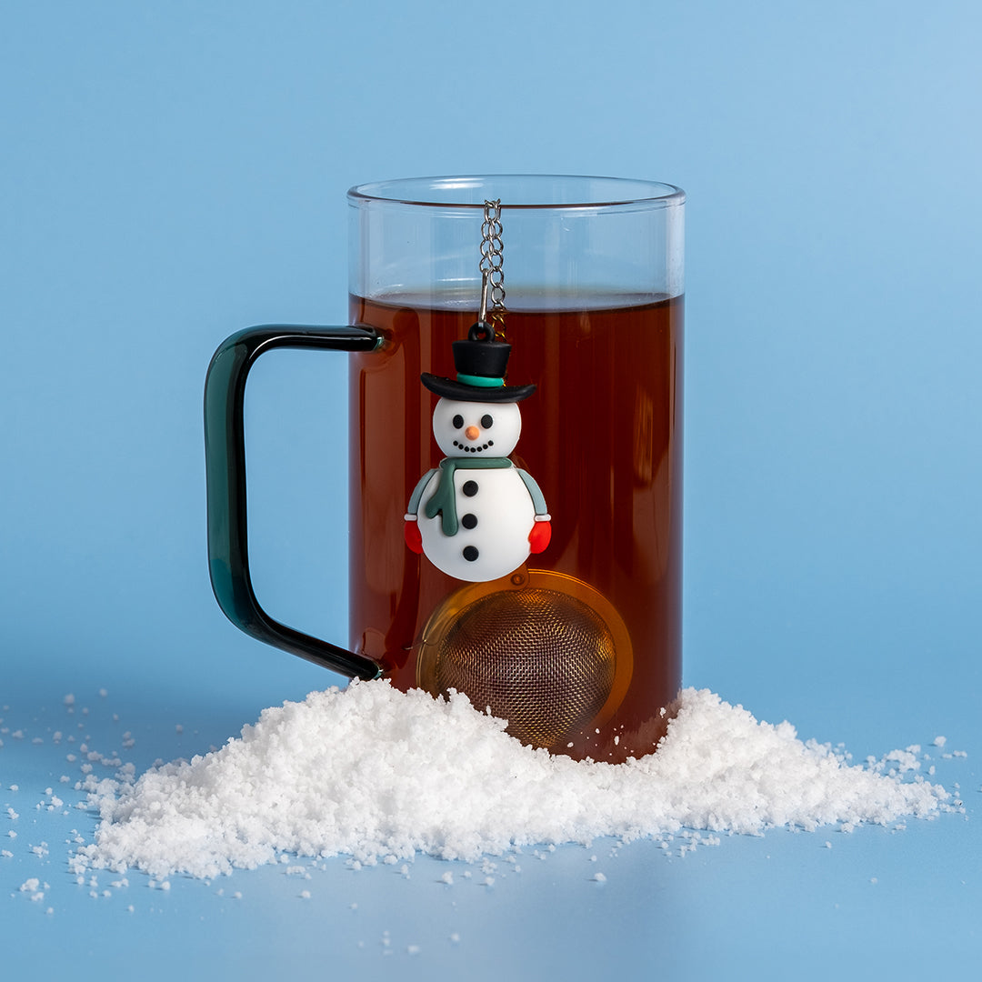 A clear glass mug filled with dark tea, featuring a hanging snowman-shaped tea infuser and a pile of snow, set against a solid light blue background.