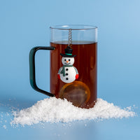 A clear glass mug filled with dark tea, featuring a hanging snowman-shaped tea infuser and a pile of snow, set against a solid light blue background.