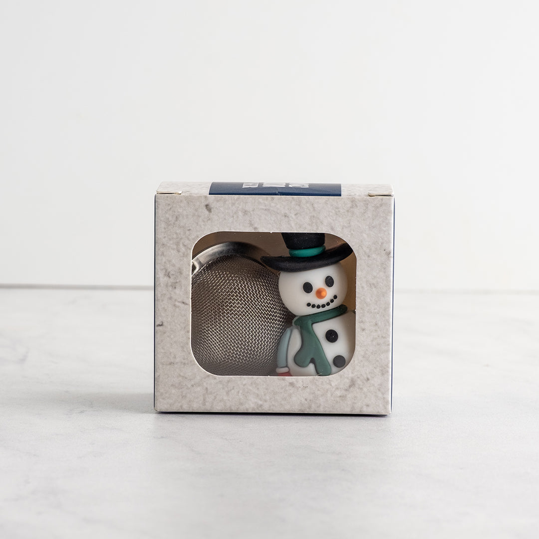 A small snowman tea infuser wearing a black top hat and green scarf next to a silver mesh ball, displayed inside a light gray textured square box with a clear window.