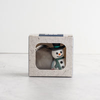 A small snowman tea infuser wearing a black top hat and green scarf next to a silver mesh ball, displayed inside a light gray textured square box with a clear window.