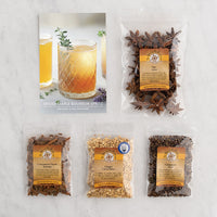 A flat-lay product arrangement on a light marble surface showing a printed recipe card for Spiced Maple Bourbon Spritz alongside five clear plastic bags of: star anise, cinnamon chunks, ginger, and cardamom seed.