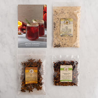 Four spice and tea products arranged on a light marble surface: a printed recipe card for spiced sangria, a bag of tea, a bag of star anise, and a bag of cinnamon plum herbal tea. The recipe card shows two red spiced drinks.