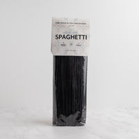 A package of black spaghetti pasta standing upright on a light gray surface with a white background. The paper label reads 'The Spice & Tea Exchange' and 'SQUID INK SPAGHETTI'.