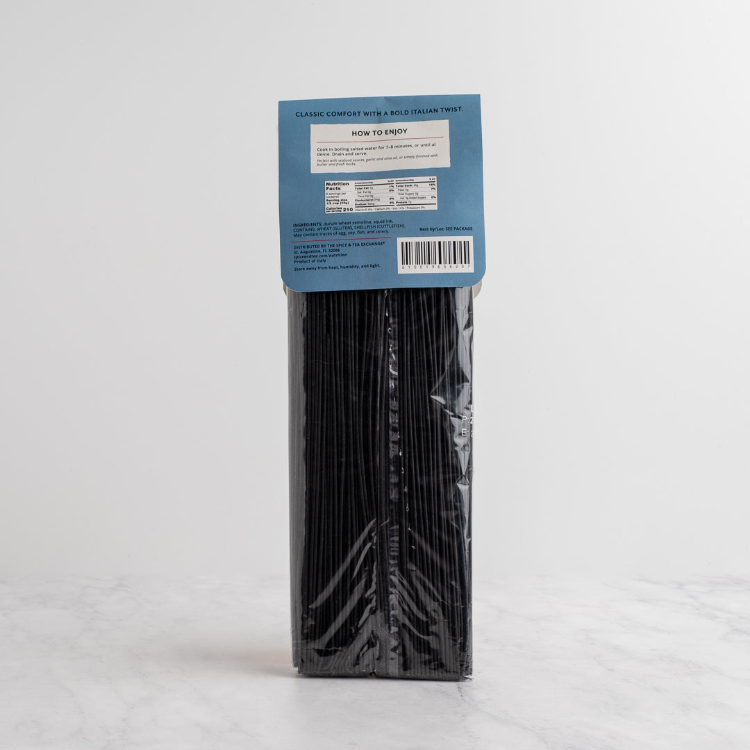 A package of black ink spaghetti from The Spice & Tea Exchange stands upright on a light marble surface against a white hexagonal tile wall. Back side.