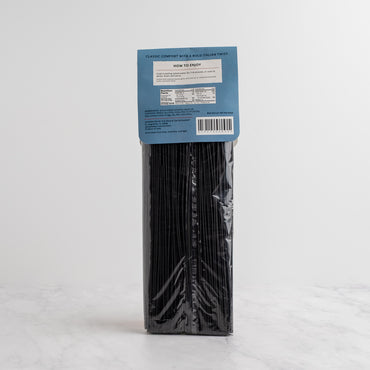 A package of black ink spaghetti from The Spice & Tea Exchange stands upright on a light marble surface against a white hexagonal tile wall. Back side.