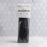 A package of black squid ink spaghetti from The Spice & Tea Exchange stands upright on a light marble surface against a white hexagonal tile wall.