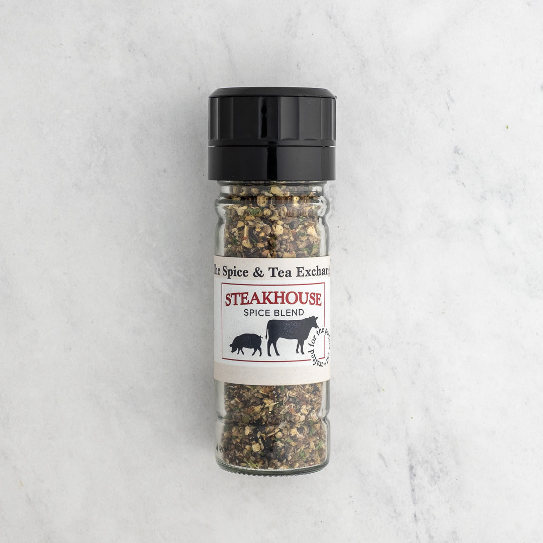 A clear glass spice jar with a black plastic grinder cap and a white paper label reading 'Steakhouse Spice Blend' from 'The Spice & Tea Exchange,' filled with a coarse spice mix, photographed on a light marble surface.