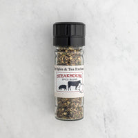 A clear glass spice jar with a black plastic grinder cap and a white paper label reading 'Steakhouse Spice Blend' from 'The Spice & Tea Exchange,' filled with a coarse spice mix, photographed on a light marble surface.