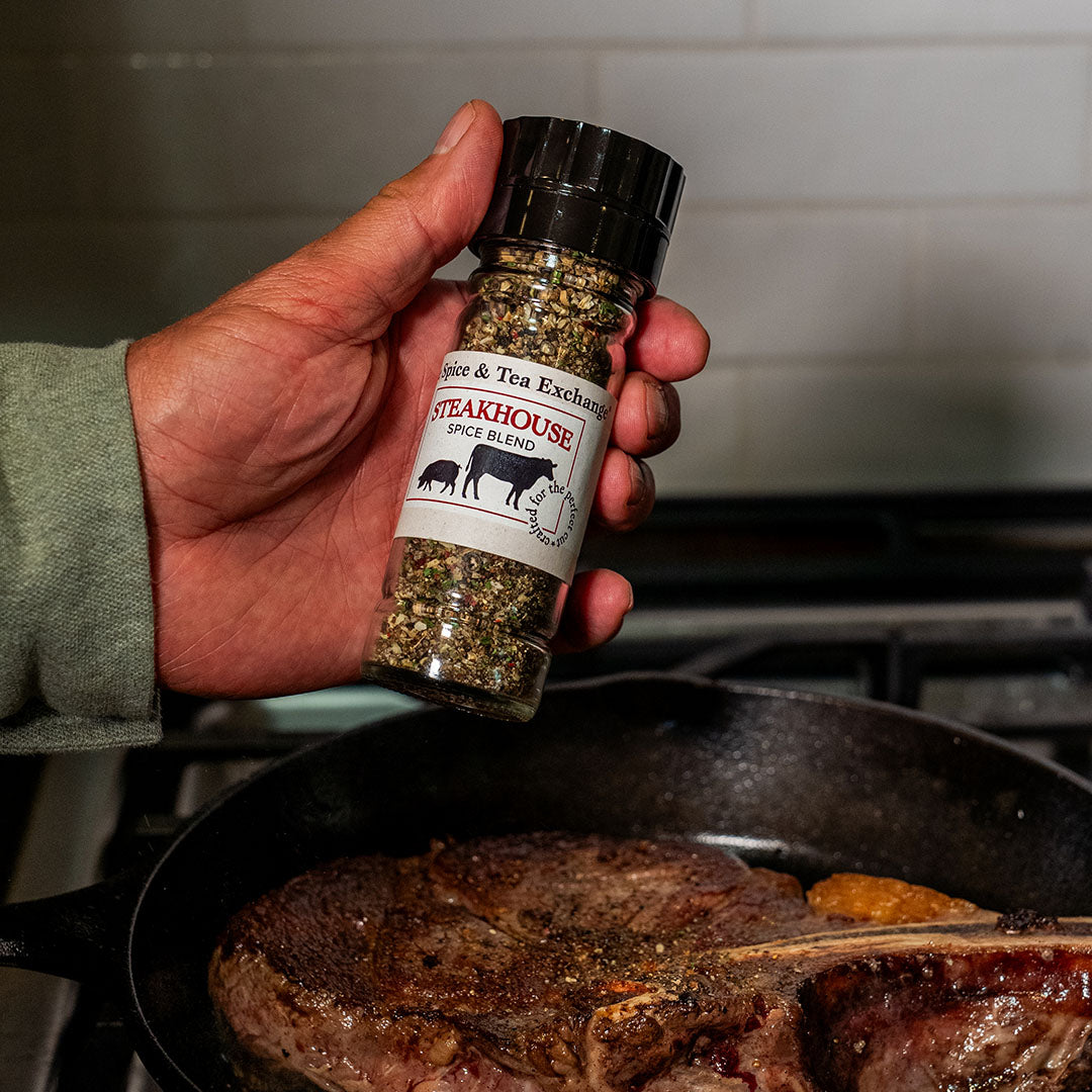 A hand holds a bottle of Steakhouse Spice Blend seasoning above a cast iron skillet with a steak cooking on a stove, with a white tiled kitchen wall in the background.